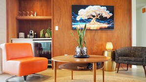 A modern living room with a round wooden coffee table, a sofa, and a chair. There is a painting of a tree on the wall, a small potted plant, and a lamp on a side table. - Tempe Pool House - Walk to ASU, Midcentury Wonder! (Tempe)