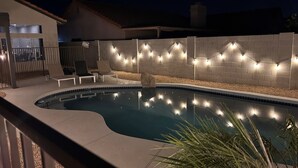 A swimming pool with a curved edge, surrounded by a pebble bed and a low brick wall. There are lounge chairs and string lights along the wall.