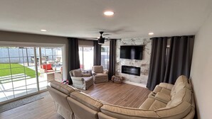 A spacious living room with a large sectional sofa, a flat-screen TV mounted on the wall, and a ceiling fan.