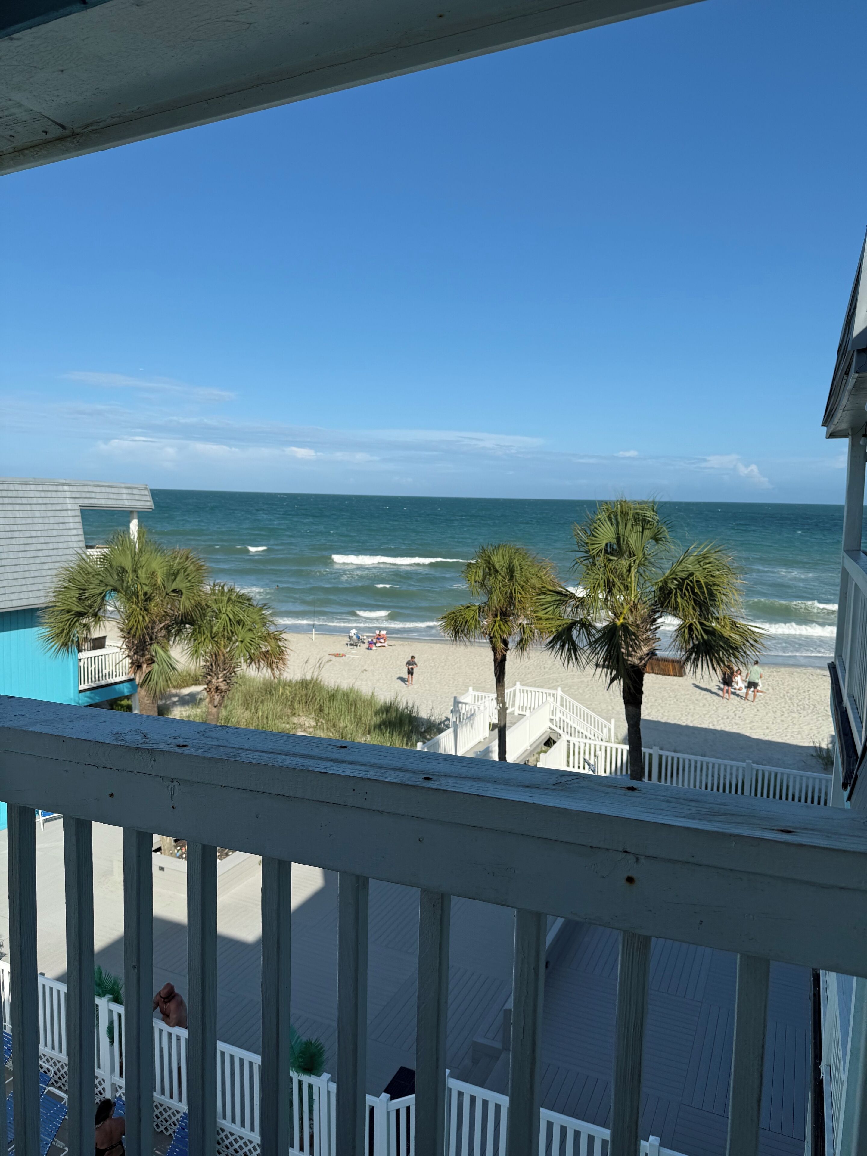 A beach view from a balcony with a wooden railing, palm trees, and a clear sky.