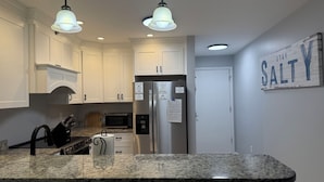 A kitchen with white cabinets, a granite countertop, a stainless steel refrigerator, and two pendant lights. - Beach House at Garden City. 2-BR condo with private pool, on the beach. (Murrells Inlet)