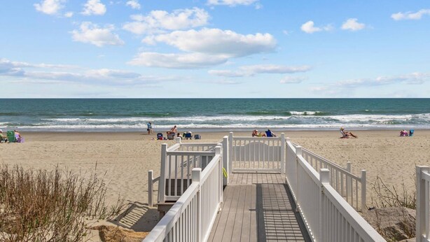 Ein hölzerner Steg führt zu einem Sandstrand mit Blick auf das Meer und einen klaren Himmel.