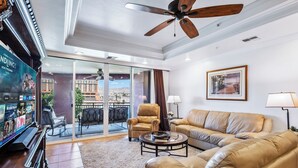 A spacious living room with a large sofa, a coffee table, and a TV. There is a ceiling fan, a framed picture on the wall, and a view of the outside through large windows. -  3 BDR Riverfront Resort w/pool, spa, dock & launch! (Bullhead City)
