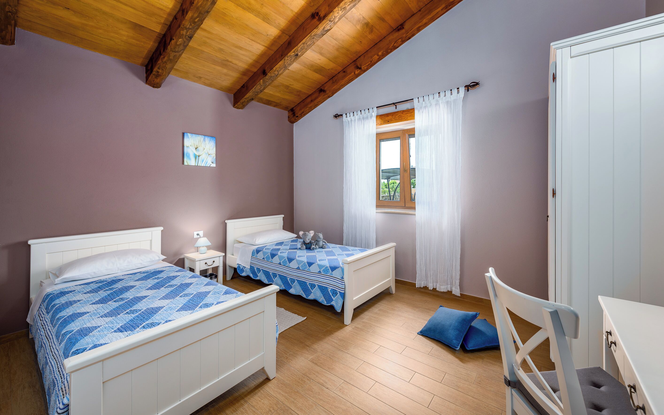 A bedroom with two beds, a wooden ceiling, a window with curtains, and a white wardrobe.