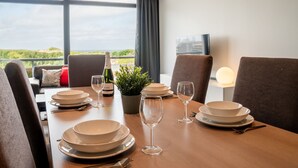 Una mesa de comedor dispuesta con platos, vasos y una botella de vino, con vistas a zonas verdes y un paisaje urbano a través de amplios ventanales.