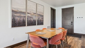 Un coin repas avec une table en bois, des chaises orange et trois grandes peintures représentant des paysages accrochées au mur.