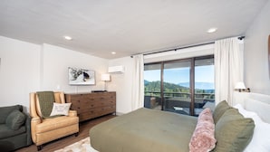 Un dormitorio con una cama grande, un aparador de madera, un sillón de cuero y vistas al exterior a través de un ventanal corredero.