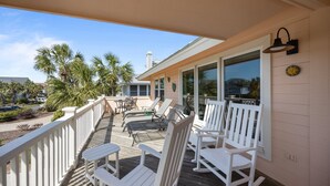 A covered outdoor deck with white rocking chairs and lounge chairs, overlooking a palm tree-lined street.