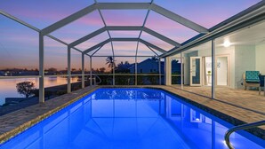 A swimming pool with a glass enclosure, a view of a lake, and a building with a door and a chair.