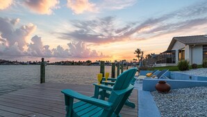 A wooden deck with chairs and a table, overlooking a body of water with a sunset in the background.