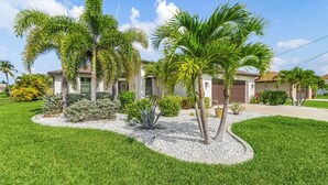 A residential house with a well-maintained lawn, palm trees, and a driveway leading to a garage. - Canal Views & Private Pool - Nirvana on Niagara - Roelens Vacations (Cape Coral)