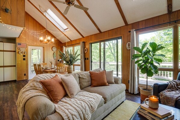 A cozy living room with a large sofa, a dining area, and a view of the outdoors. - FALL HIDEOUT 3BR RETREAT•BIG DECK,GAME RM, FIREPIT,FIREPLACE•6BIKES•GOLF•FARMS (Lake Ariel)