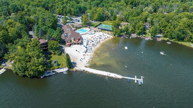 Eine Luftaufnahme eines See-Resorts mit Strand, einer Anlegestelle und einem Poolbereich, umgeben von dichtem Wald.