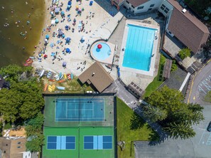 An aerial view of a property with a swimming pool, tennis courts, and a beach area with umbrellas.