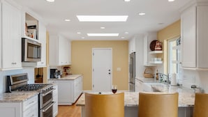 A modern kitchen with white cabinetry, a built-in oven, a microwave, and a gas stove. There are two bar stools with a wine glass on one.