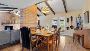 A dining area with a wooden table set for four, surrounded by wooden chairs. A brick wall and a fireplace are visible in the background.