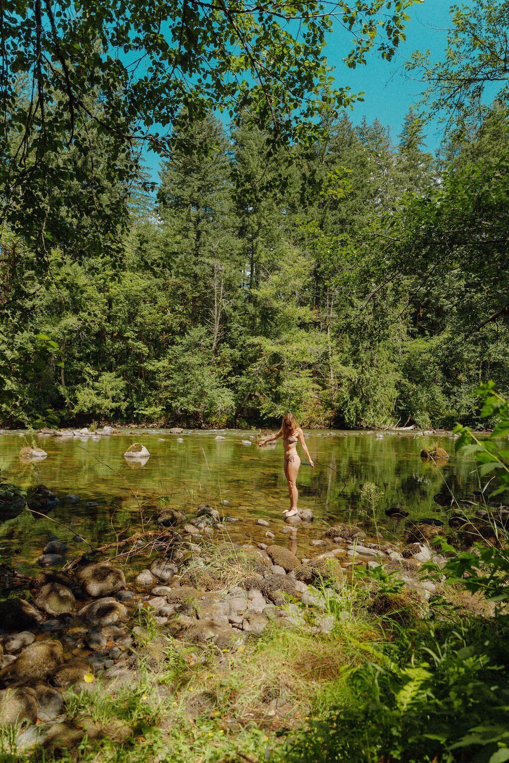 A woman standing by a clear river surrounded by dense forest.