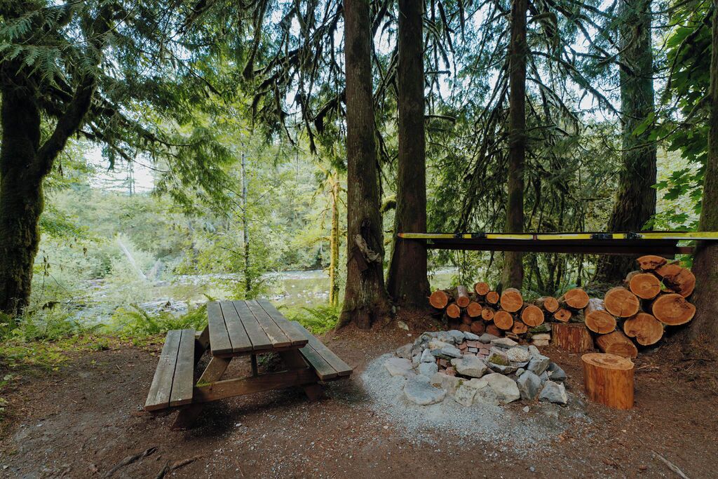 En campingplats i ett skogsområde med ett picknickbord, en eldstad och staplade vedträn.