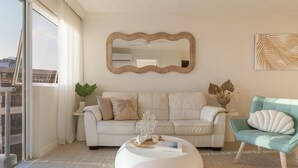A modern living room with a white sofa, a round white coffee table, and a teal armchair. A large mirror hangs on the wall, and there is a potted plant on the side table. - 1BR Beachfront Retreat ~ Azure Beachfront Bliss (Palm Beach)