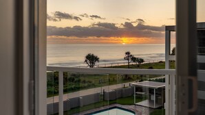 Vista da un balcone su una piscina, un'area verde e un tramonto sull'oceano.