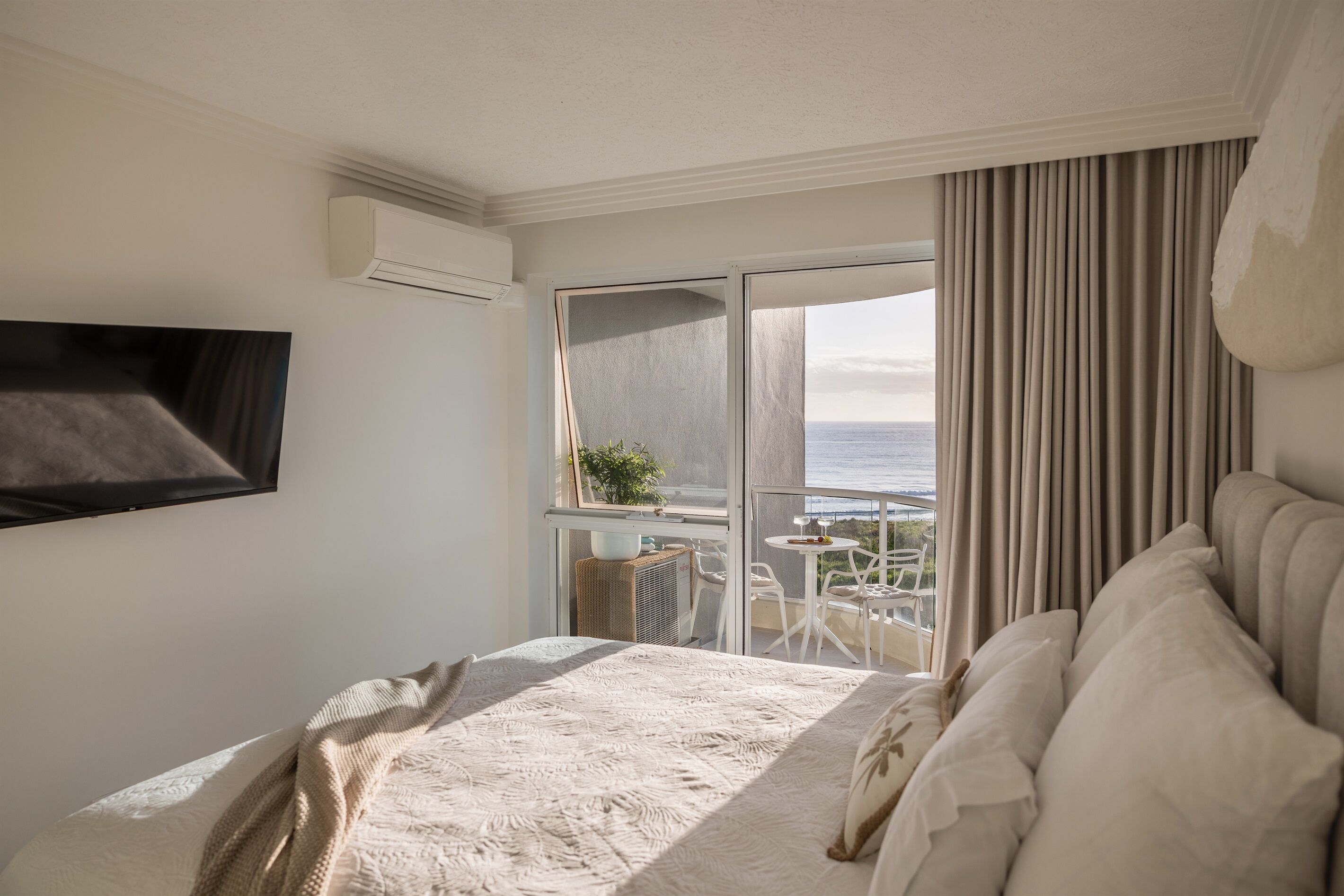 A hotel room with a bed, a TV, a balcony with a table and chairs, and a view of the sea.