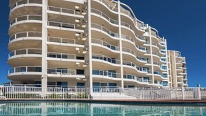 Un hotel a più piani con balconi, una piscina e un cielo azzurro intenso.