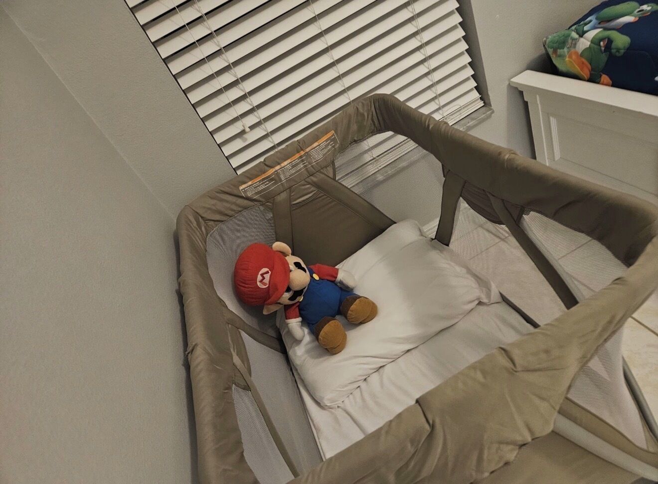 A crib with a plush toy, a window with blinds, and a white dresser.