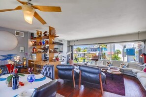 A modern living room with a dining area, a ceiling fan, and a large window with a view of buildings.