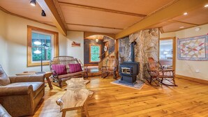 A cozy living room with a stone fireplace, wooden furniture, and large windows.