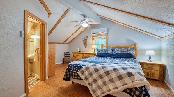 A bedroom with a sloped ceiling, a bed with a checkered blanket, a wooden nightstand, a lamp, and a ceiling fan.