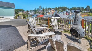 A wooden deck with Adirondack chairs and a stone grill, overlooking a residential area with colorful houses and a clear blue sky. - Sand Seeker in Olivia Beach (Lincoln City)