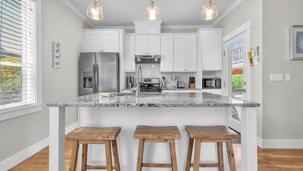 A modern kitchen with white cabinets, a granite countertop, and three wooden stools. - Sand Seeker in Olivia Beach (Lincoln City)