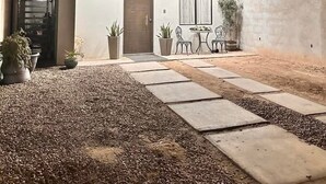 A modern two-story house with a patio area featuring a dining table and chairs, surrounded by potted plants and a gravel pathway. - Beautiful house in col independencia (Mexicali)