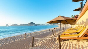 A beach with sun loungers and umbrellas, a clear sky, and a boat in the water. - Enjoy your Thanksgiving in the Sun and on the Beach Los Cabos (Cabo San Lucas)