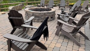 A fire pit surrounded by Adirondack chairs on a brick patio with a mountain view.