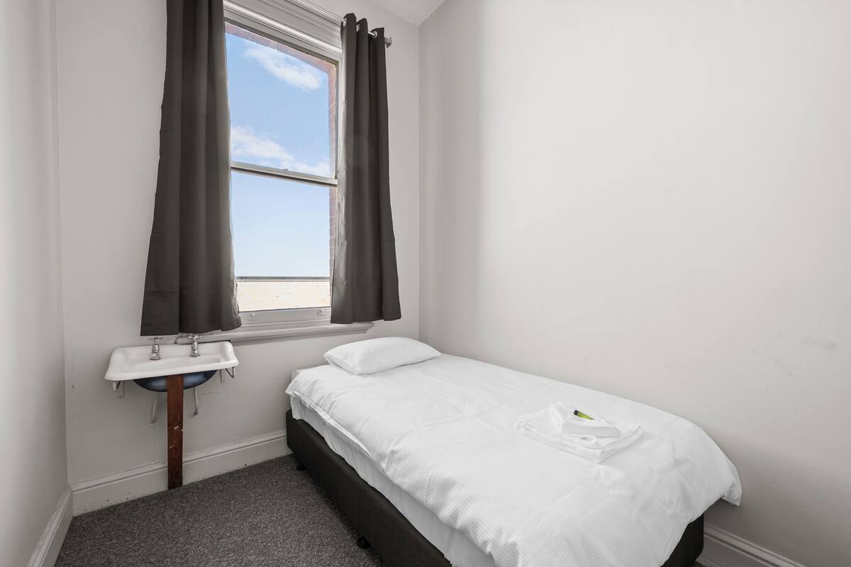Una habitación pequeña y limpia con una cama individual, una ventana con cortinas y un lavamanos.
