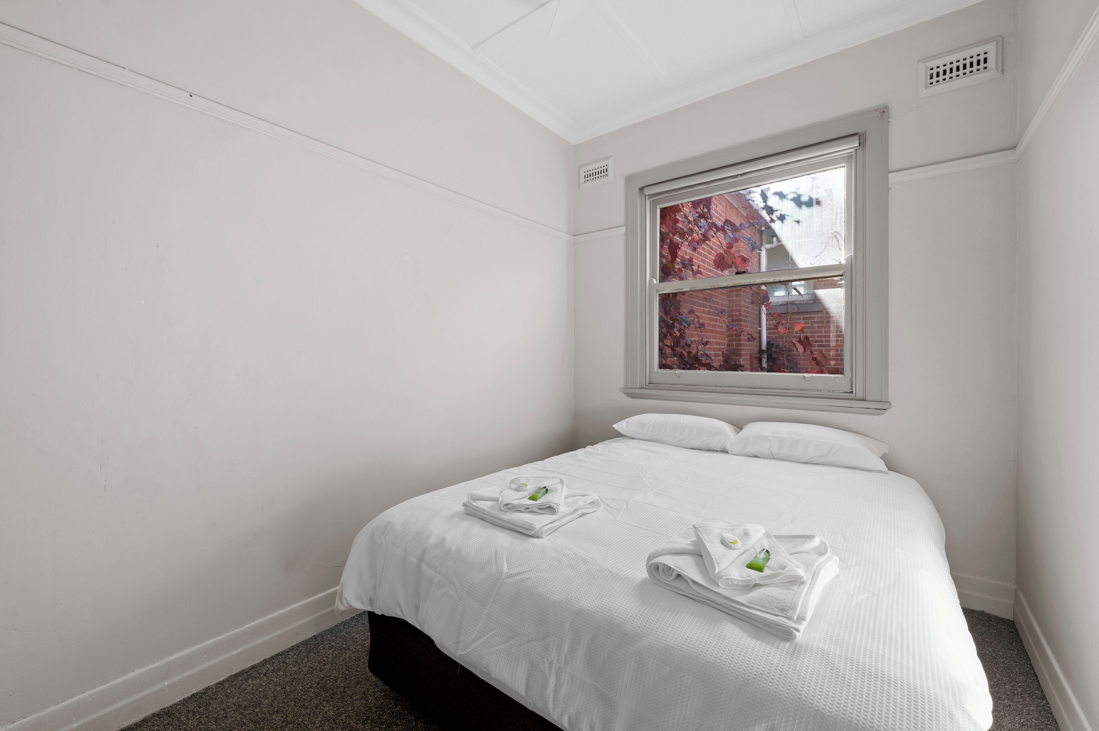A hotel room with a neatly made bed, white walls, a window with a view of a brick building, and a vent on the ceiling.