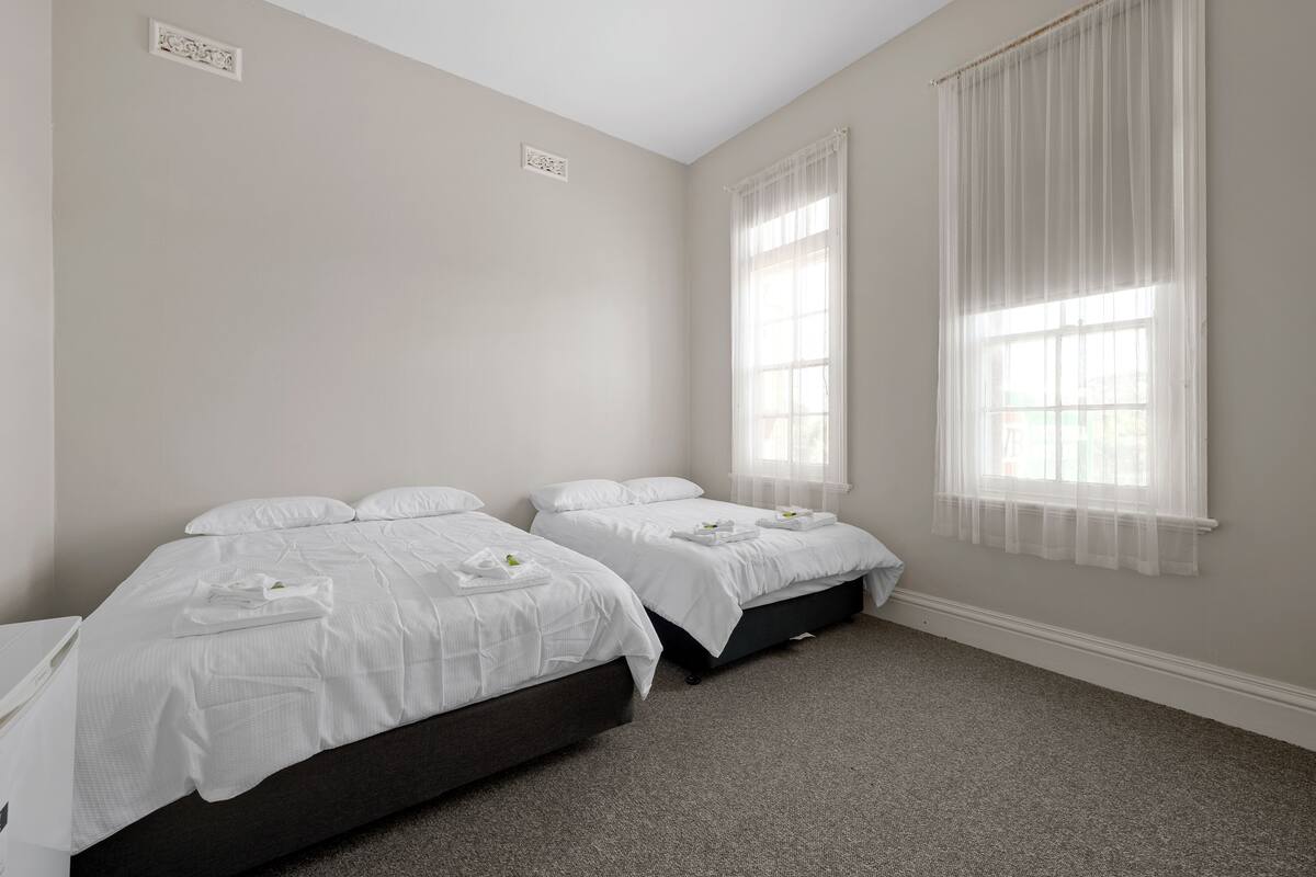 Habitación de hotel con dos camas, piso alfombrado y un ventanal con cortinas translúcidas.