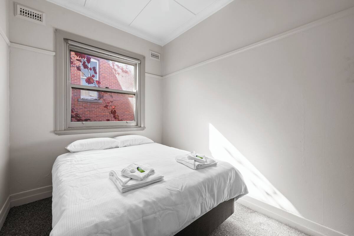 Una cama bien hecha con sábanas blancas y dos toallas dobladas, una ventana con vista a un edificio de ladrillo y un techo blanco con una rejilla.