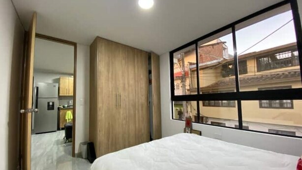 A hotel room with a bed, a large window, a wardrobe, and a view of the outside buildings.
