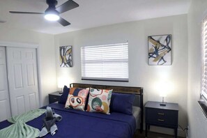 Une chambre à coucher comprenant un lit, un ventilateur de plafond, une fenêtre avec des stores et deux tableaux accrochés au mur.