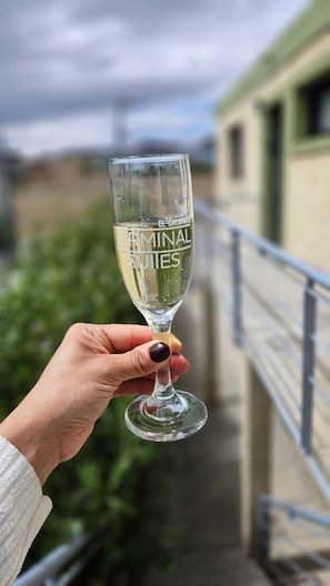 A hand holding a glass with "EL GIMLAPPA EL TERMINAL SUITES" text, overlooking a balcony with a railing and greenery.