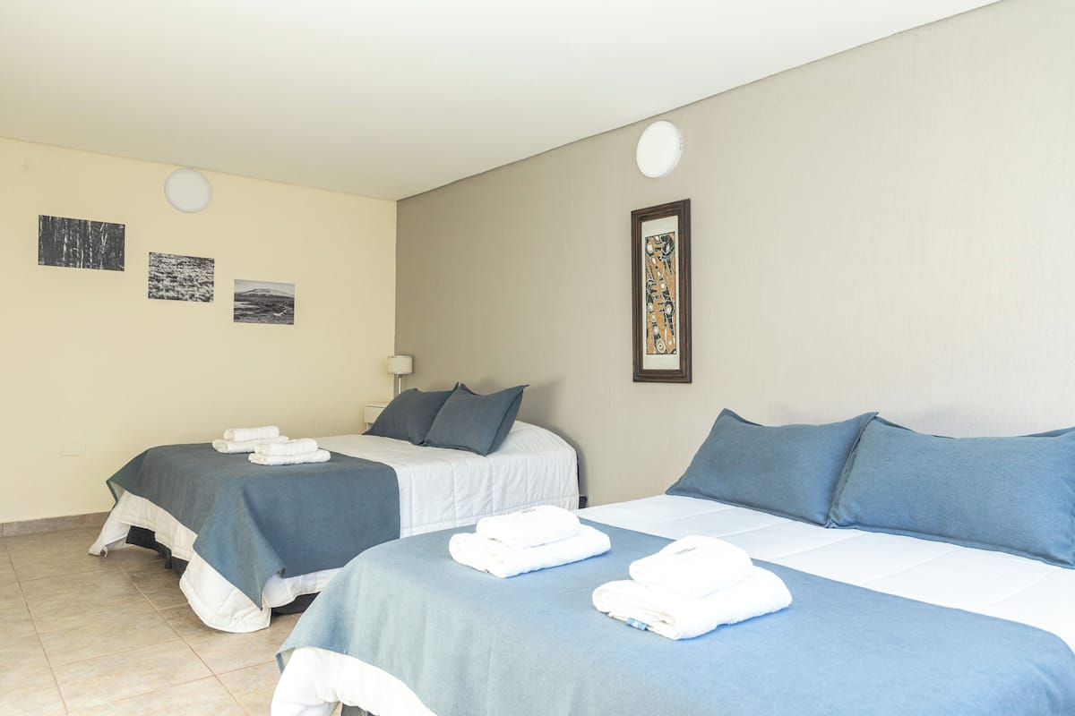 A hotel room with two beds, each with blue bedding and white towels. There are three framed pictures on the wall.