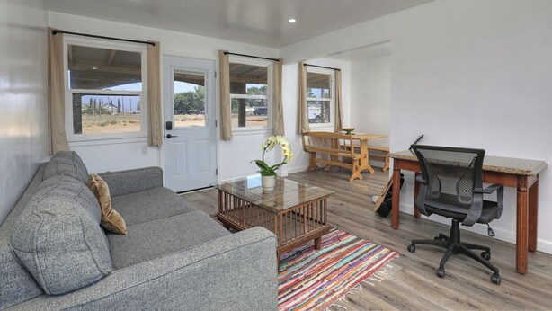 A spacious living room with a grey sofa, a wooden coffee table, a colorful rug, a wooden dining table with chairs, and a black office chair.