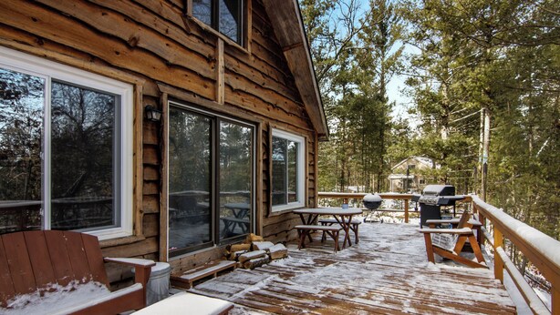 A wooden deck with outdoor furniture, a barbecue grill, and a view of a forested area. - Cozy 3-bedroom cabin on the Pere Marquette River (Baldwin)