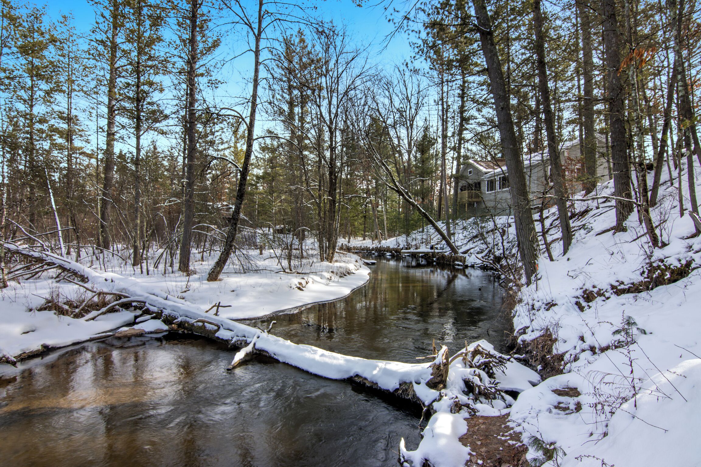 A snow-covered landscape with a stream, trees, and a building in the background.