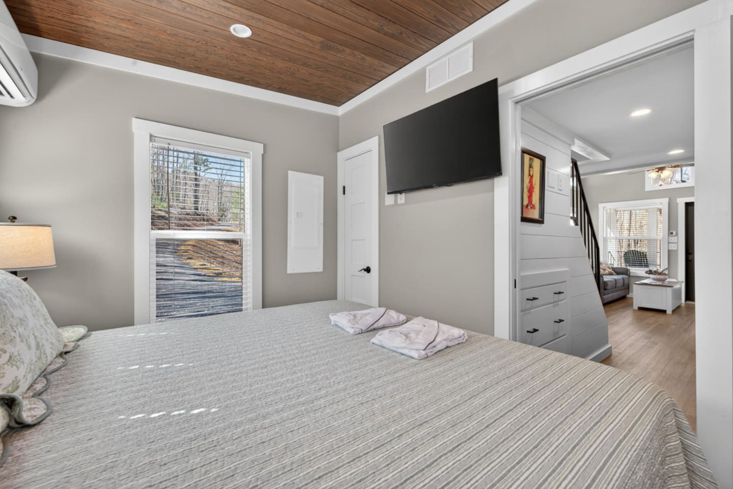 Un dormitorio con cama, televisor montado en la pared, ventana con persianas y una escalera que lleva a otra habitación.