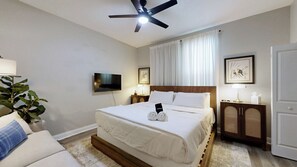 Een hotelkamer met een bed, een plafondventilator, een flatscreen-televisie, een nachtkastje met een lamp, een ingelijst schilderij en een deur.