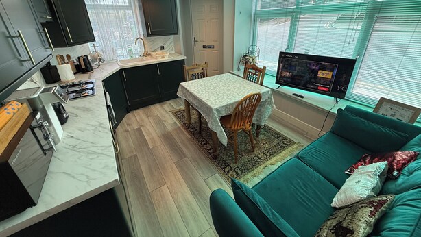 Une cuisine moderne avec des meubles sombres, un plan de travail en marbre et un four encastré. On y trouve un coin repas avec une table et des chaises, ainsi qu’un canapé turquoise agrémenté de coussins décoratifs. Un téléviseur à écran plat est fixé au mur, et une fenêtre avec des stores est visible.