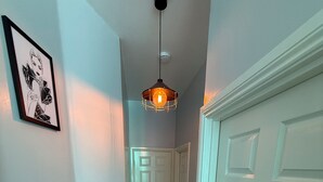 Un couloir avec un plancher en bois, une porte blanche, un panier à linge et un luminaire suspendu.